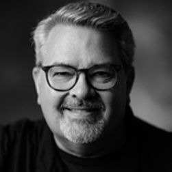 A man with glasses and a beard smiles, in a black and white portrait.