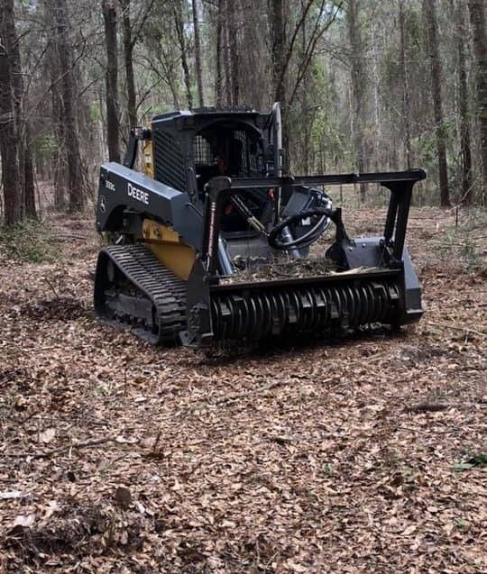 Black tracked skid steer in a wooded area, clearing leaves on a forest floor.