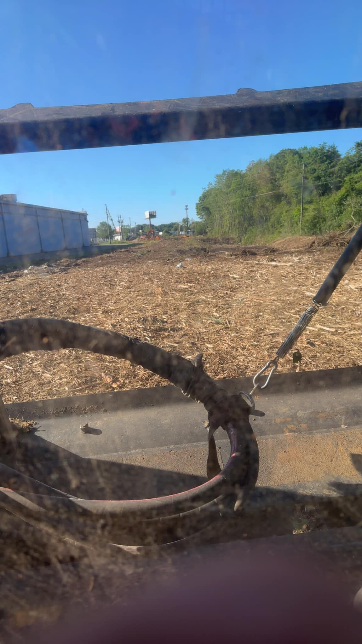View through a vehicle window of a sunlit dirt lot with piles of wood chips, trees, and buildings in the distance.