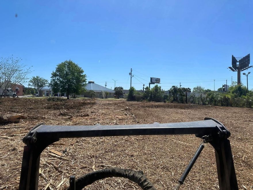 View from a bicycle over a dry dirt lot with trees, utility poles, and a clear blue sky.