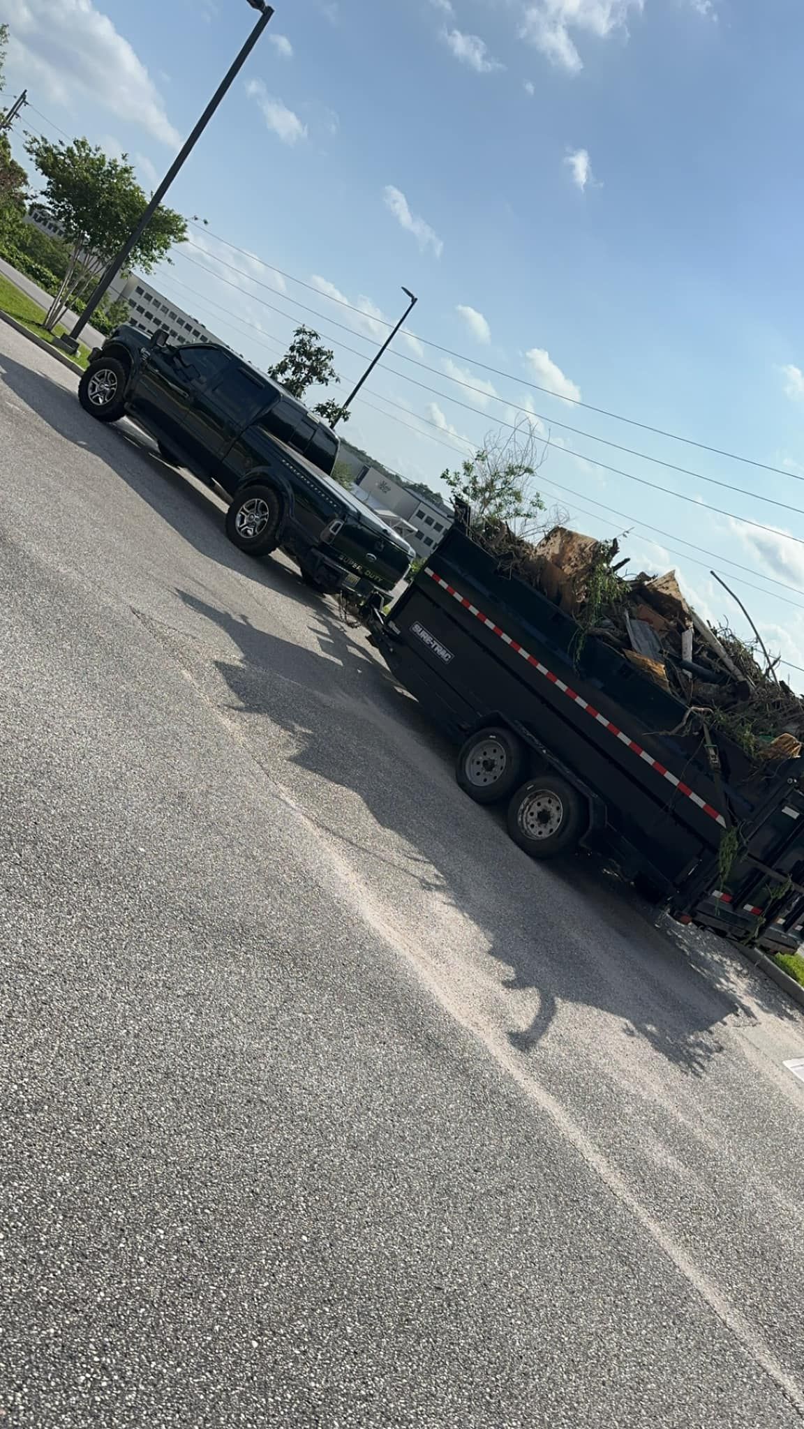 Black pickup truck towing a flatbed trailer loaded with brush on a sunny roadside.