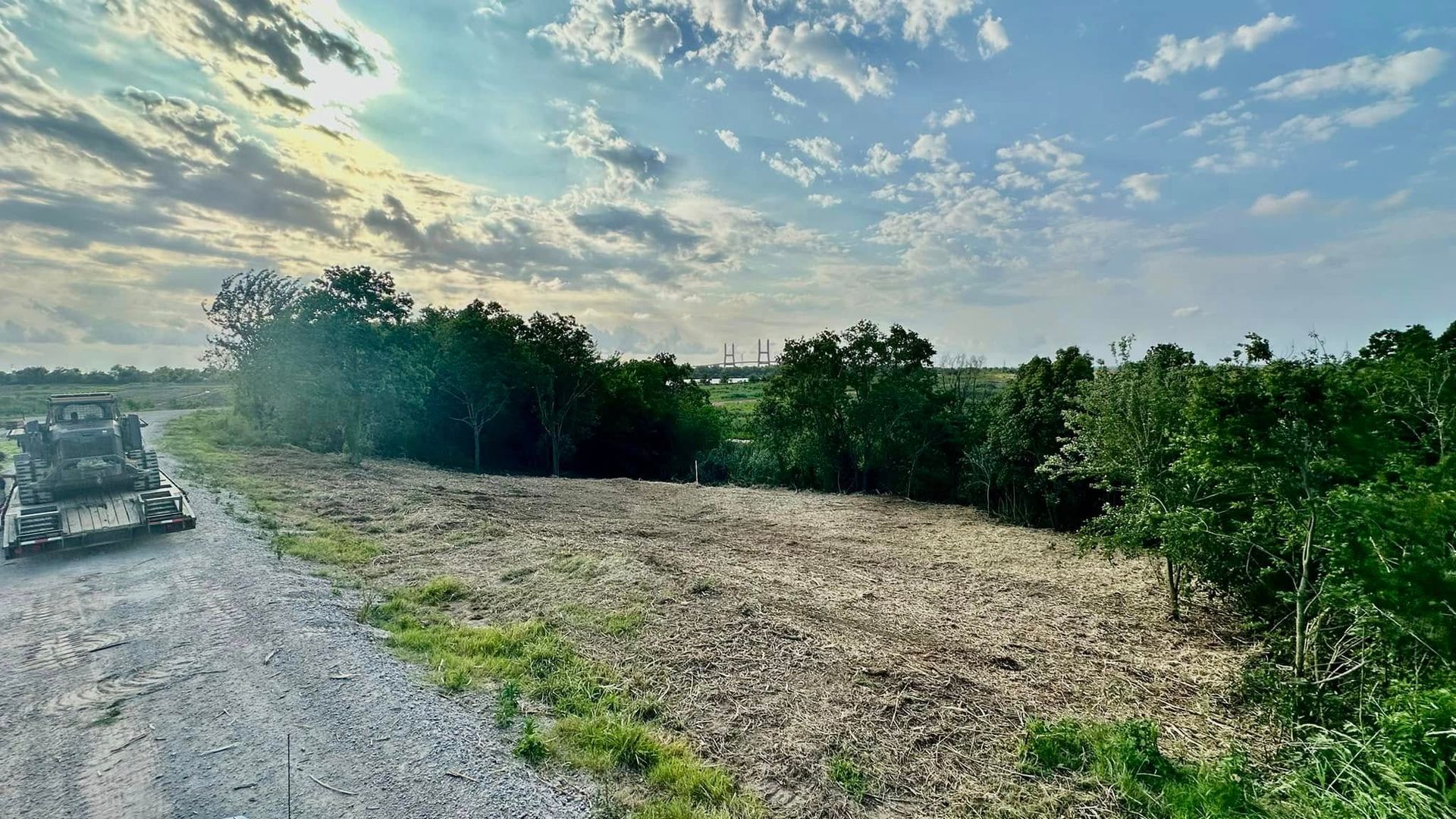 Gravel roadside beside a grassy field and trees under a cloudy blue sky