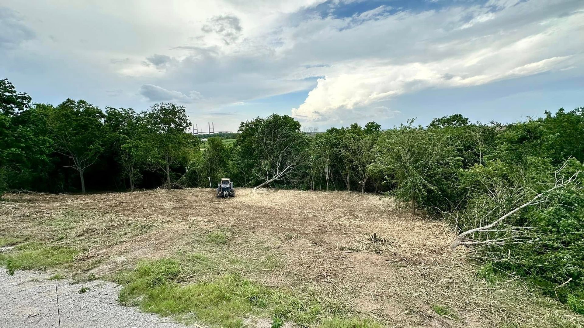 Cleared field with a small vehicle near dense green trees under a cloudy sky