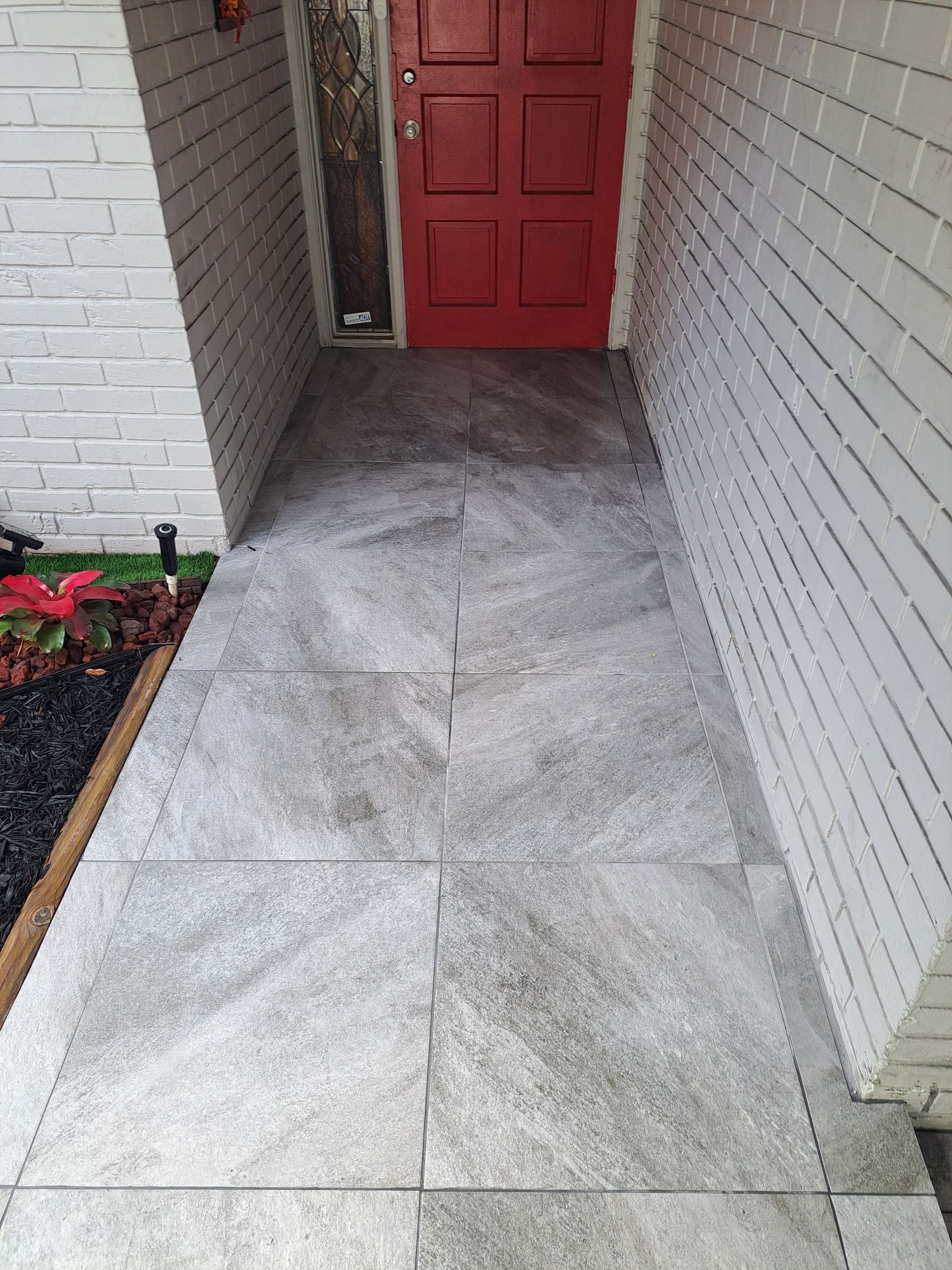 A tiled walkway leading to a red door with a white brick wall.