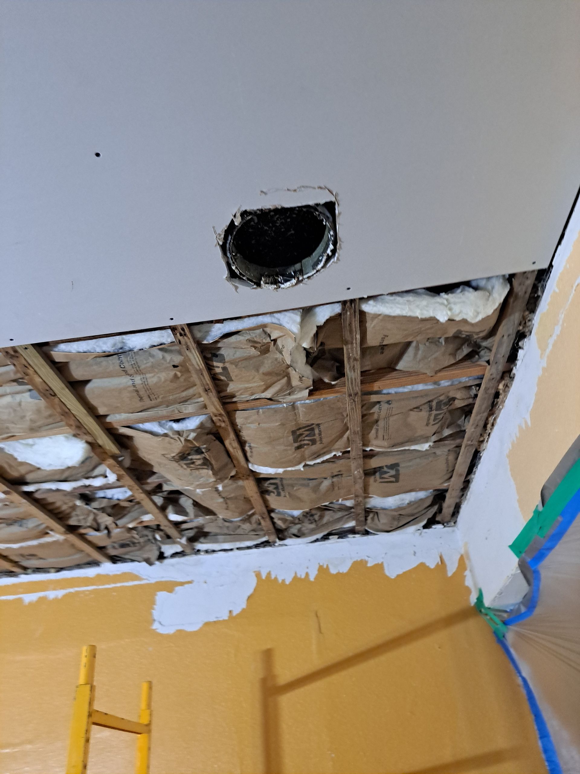 A hole in the ceiling of a room with a ladder in the background.