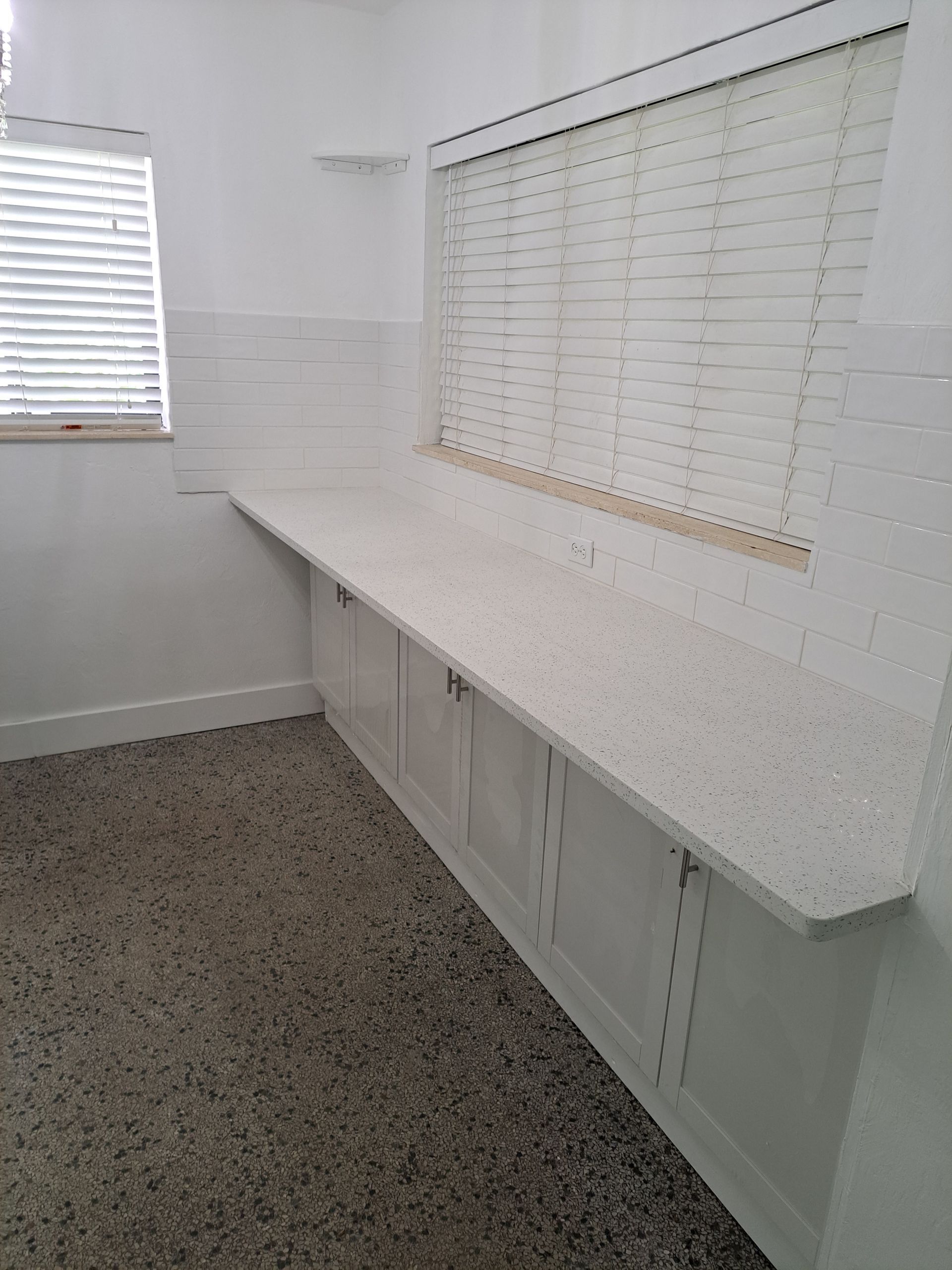 A kitchen with white cabinets , white counter tops , and a window with blinds.