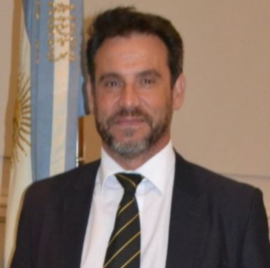 Hombre con traje y corbata, con una bandera argentina de fondo, sonriendo levemente.