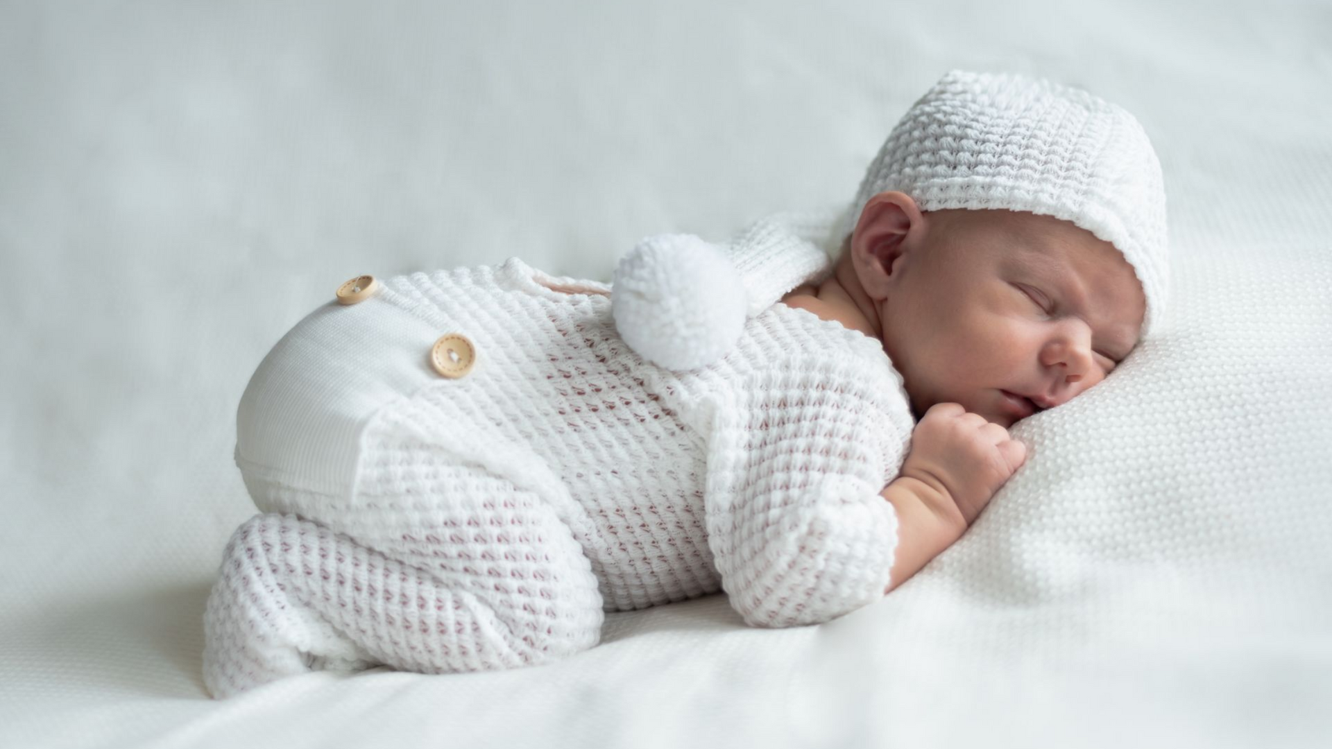 Een pasgeboren baby in een wit pakje en mutsje slaapt op een bed.