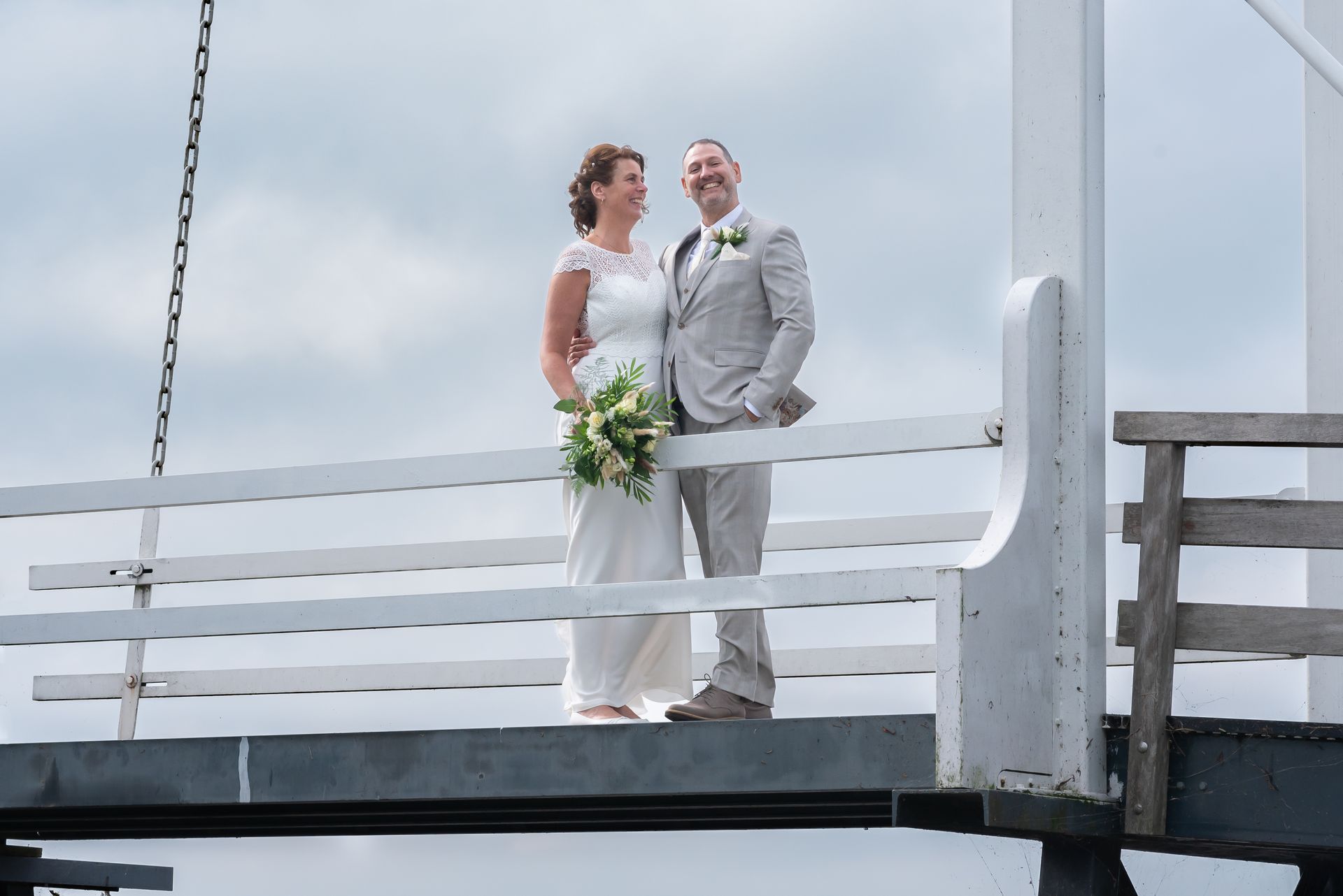 Bruid en bruidegom staan ​​op een witte brug in Nieuwkoop met bloemen in hun handen, onder een bewolkte hemel.
