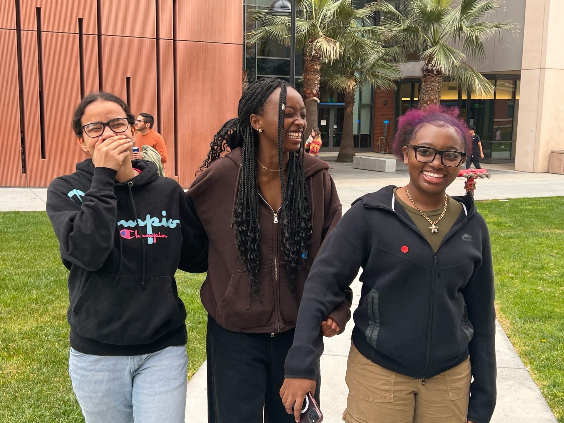 Three students are walking down a sidewalk holding hands and smiling.