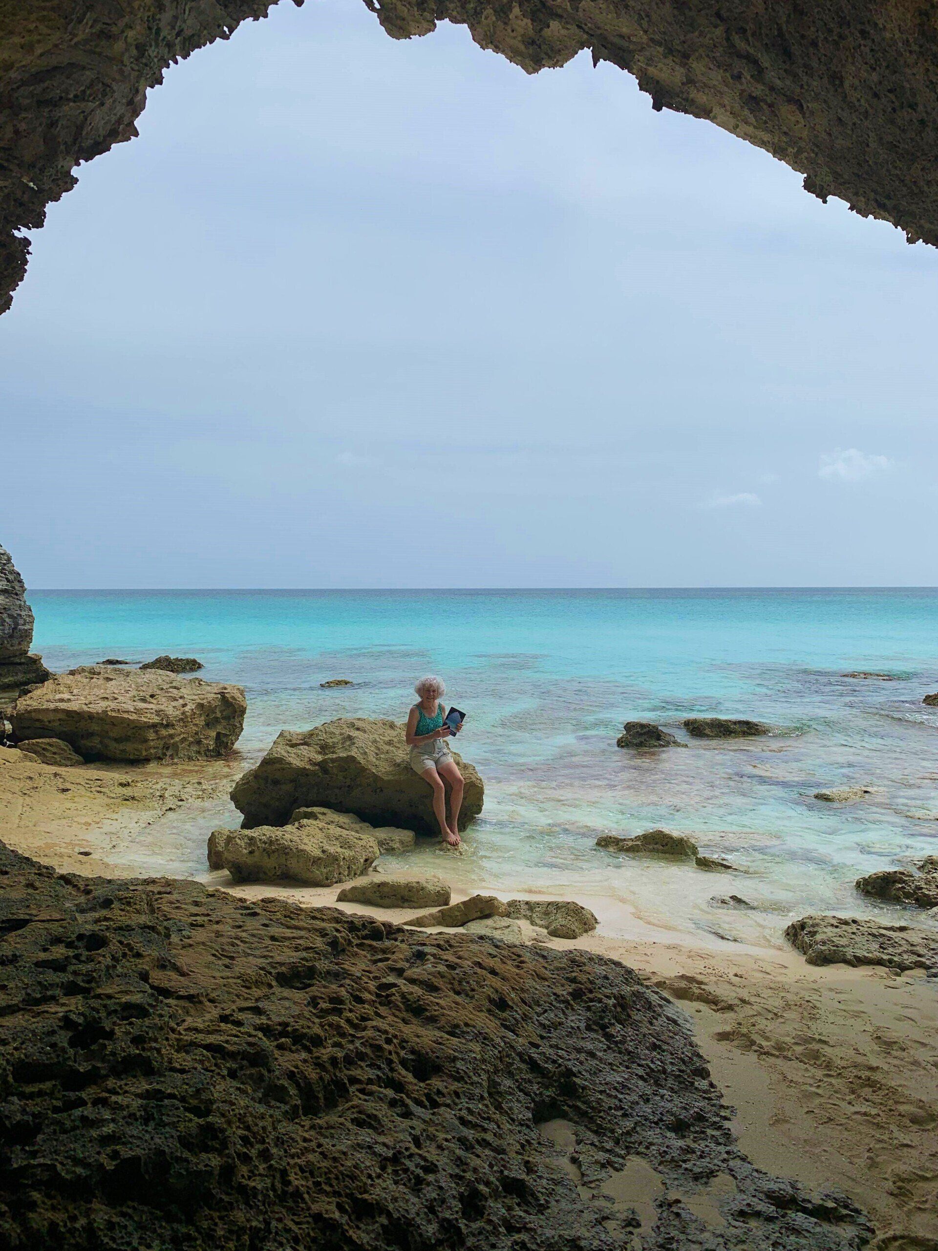 A woman is sitting on a rock near the ocean outside of the cave that features on the cover of uncharted a novel by janet howle.
