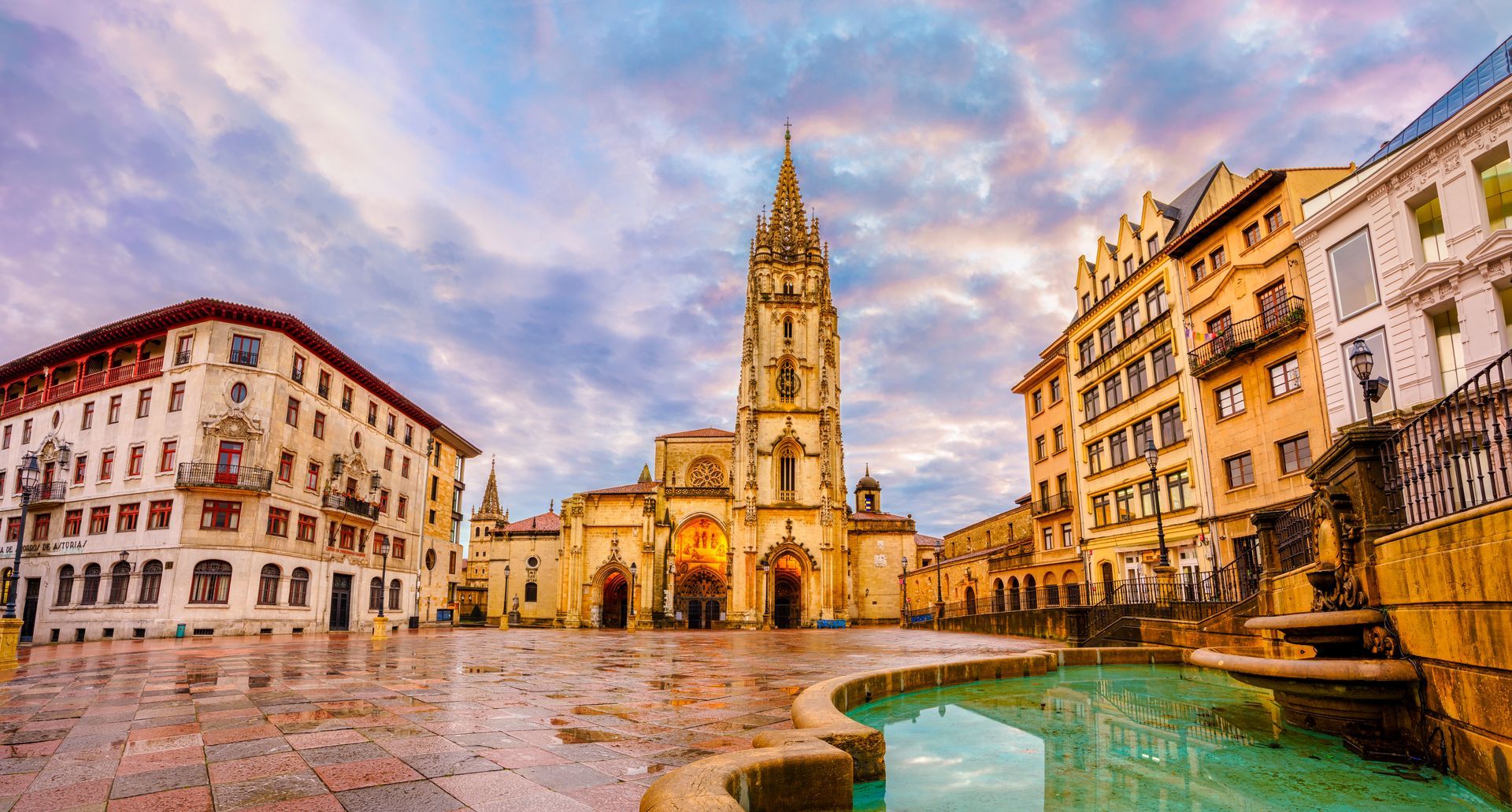 A city square with a clock tower and a fountain in the middle of it.