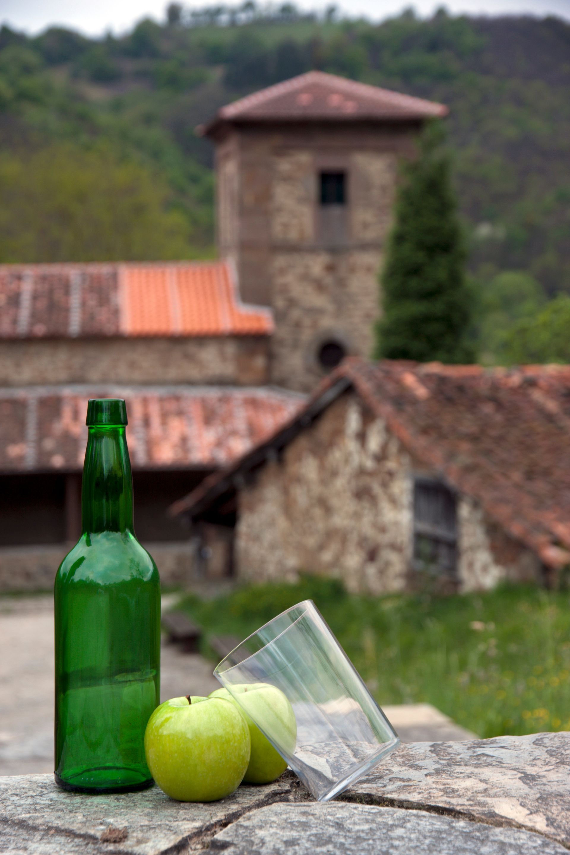 A bottle of wine , two apples and a glass are on a stone wall.