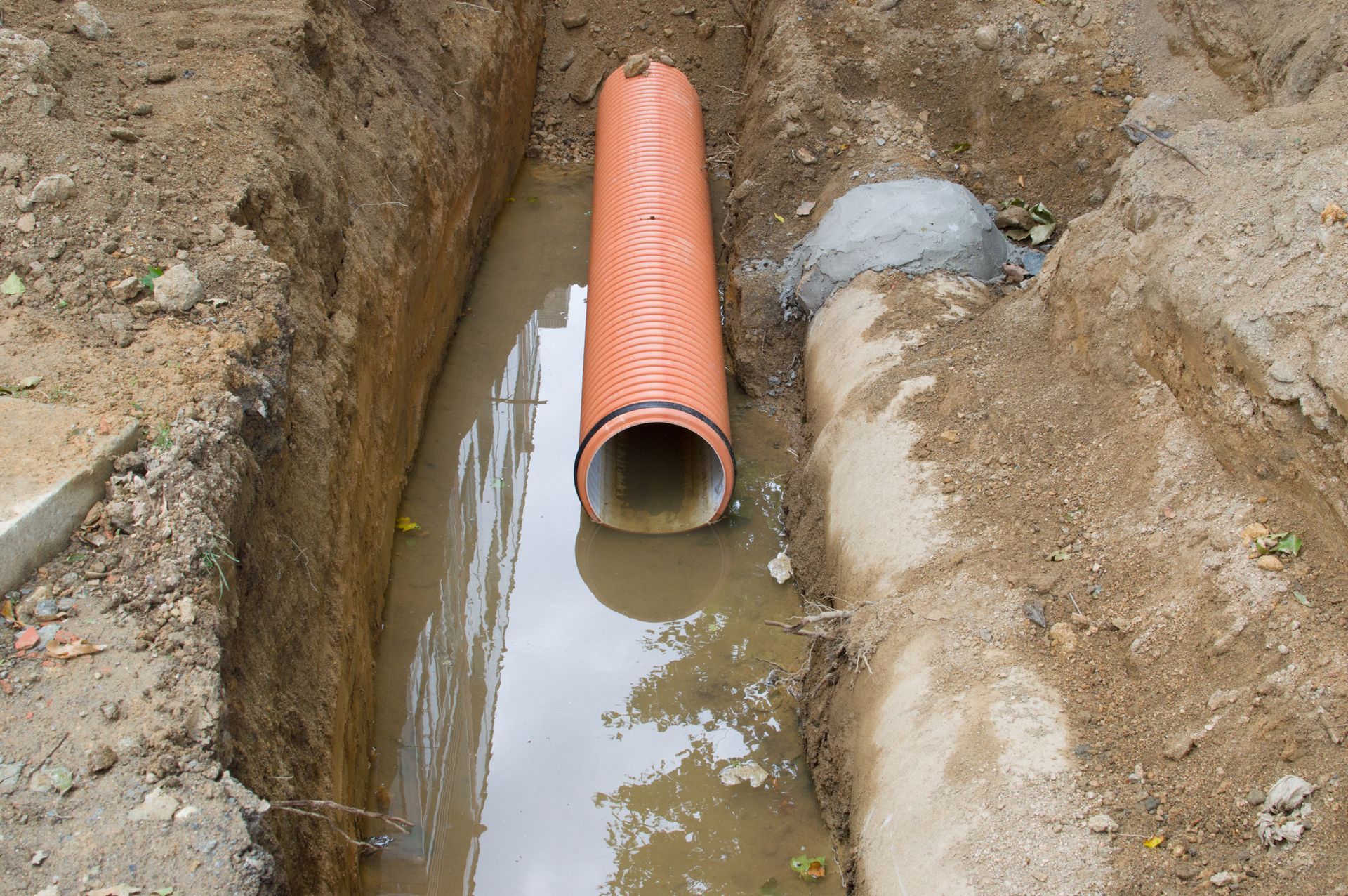 An orange pipe is laying in the dirt next to a concrete pipe