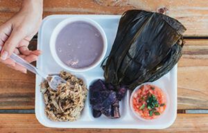 A person is eating food from a styrofoam tray on a wooden table.