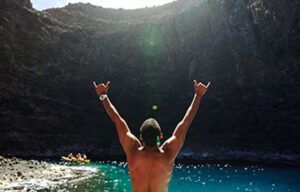 A shirtless man is standing in the water with his arms in the air.