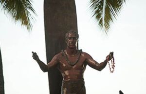 A statue of a man standing next to a surfboard holding a lei.