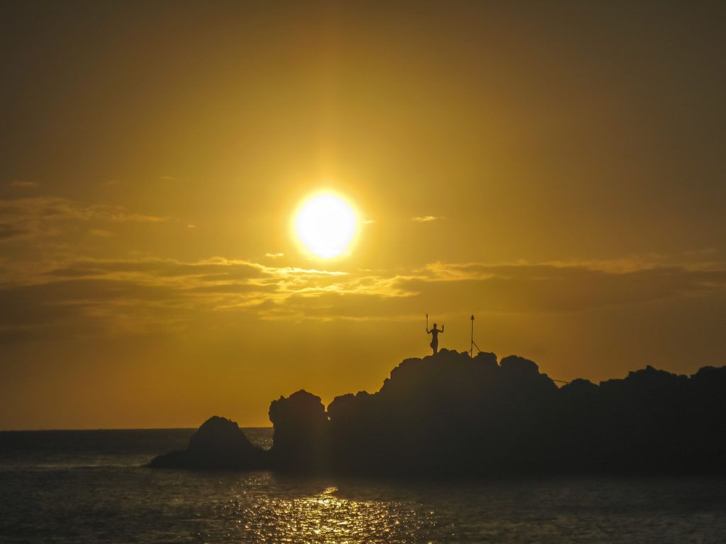 A sunset over the ocean with a person standing on a rock in the foreground.