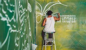 A man is sitting on a ladder painting a mural on a wall.