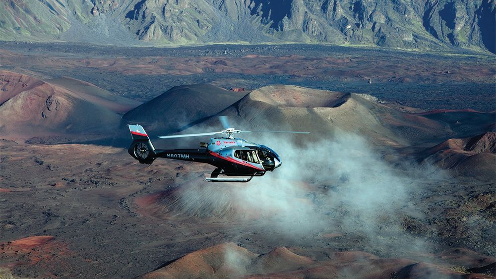 A helicopter is flying over a desert landscape with mountains in the background.