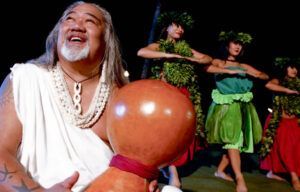 A man is holding a gourd in front of a group of hula dancers.