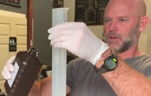 A man wearing gloves is holding a measuring cup and a bottle.