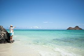 A man is standing on a rock on a beach looking at the ocean.