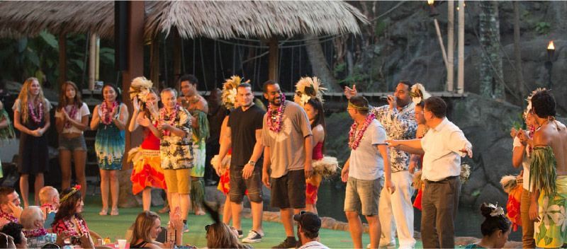A group of people are dancing in front of a thatched hut.