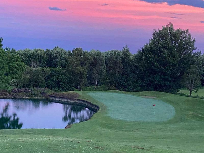 A golf course with a pond in the middle of it and a sunset in the background.