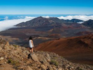 A woman is standing on top of a mountain looking at the clouds.