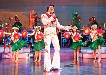 A man in a hawaiian suit is singing into a microphone on a stage surrounded by hula dancers.