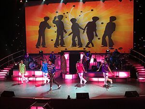 A group of people are dancing on a stage in front of a large screen.