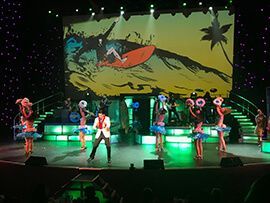 A group of people are dancing on a stage in front of a large screen.
