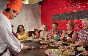A group of people are sitting at a table in a restaurant while a chef prepares food.