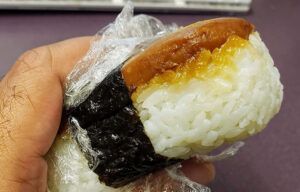 A person is holding a piece of sushi wrapped in plastic wrap.