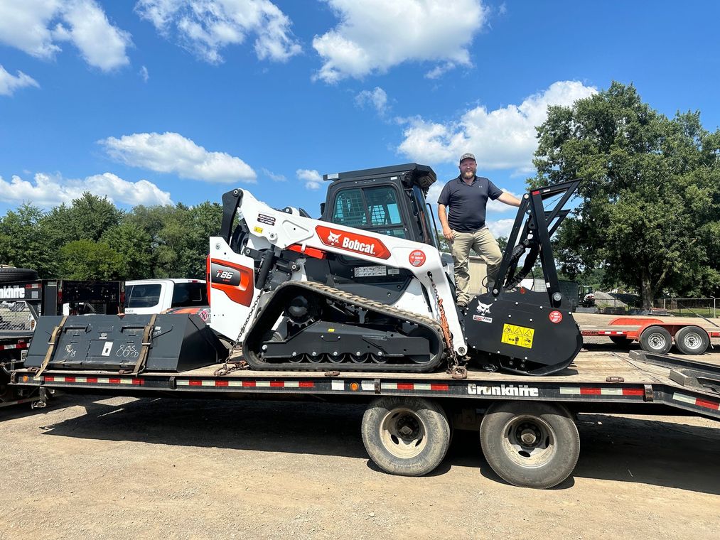 Man standing beside a compact loader on a trailer under a blue sky
