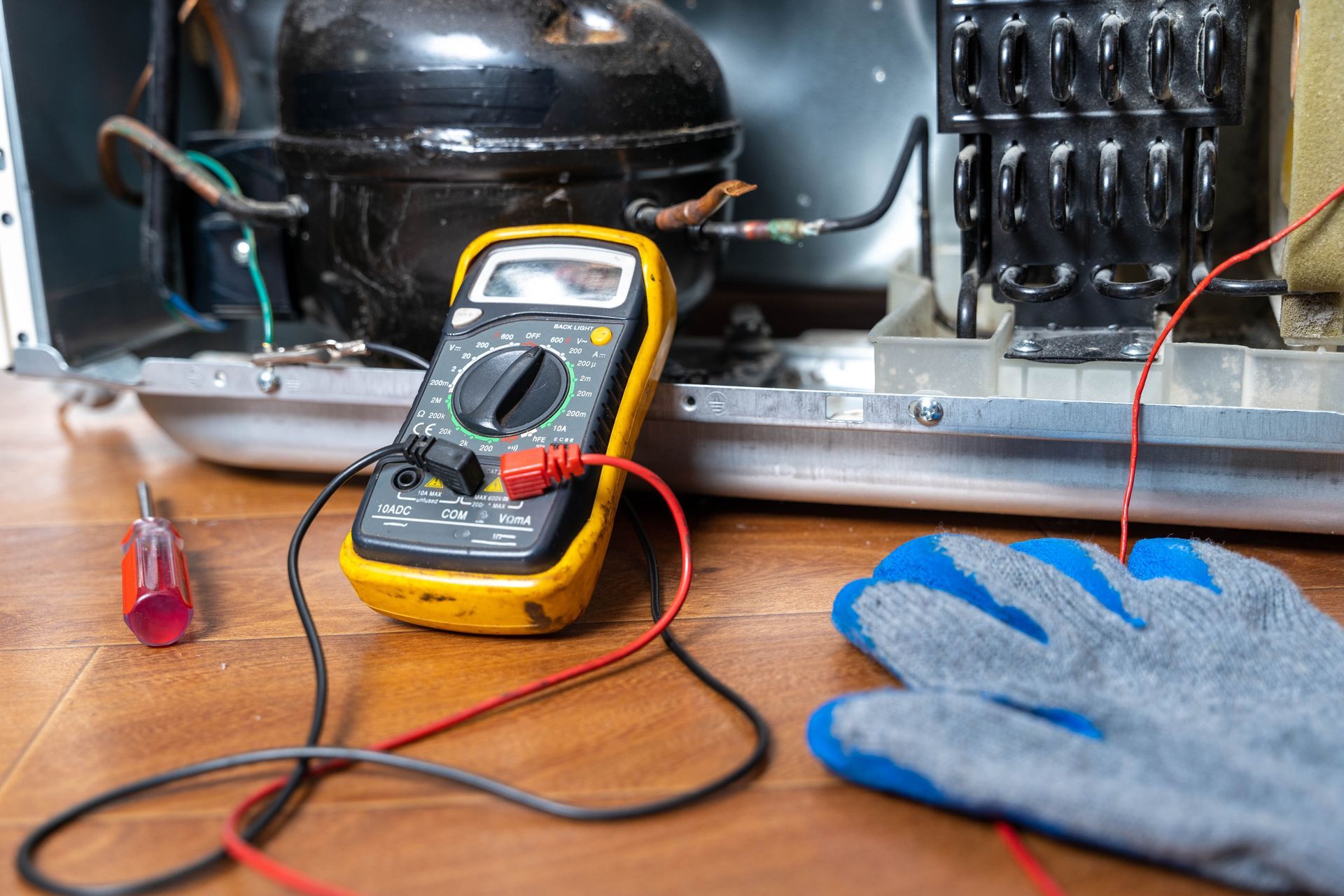 Multimeter testing wires on a kitchen appliance on a wooden counter, with a screwdriver and work glove nearby