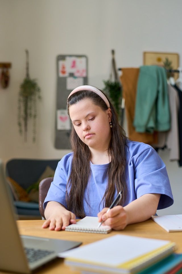 A young woman with down syndrome is sitting at a table writing in a notebook.