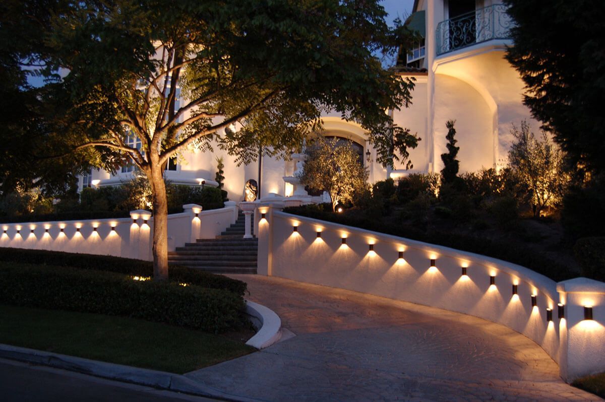 Illuminated driveway and retaining wall leading to a white mansion at night.