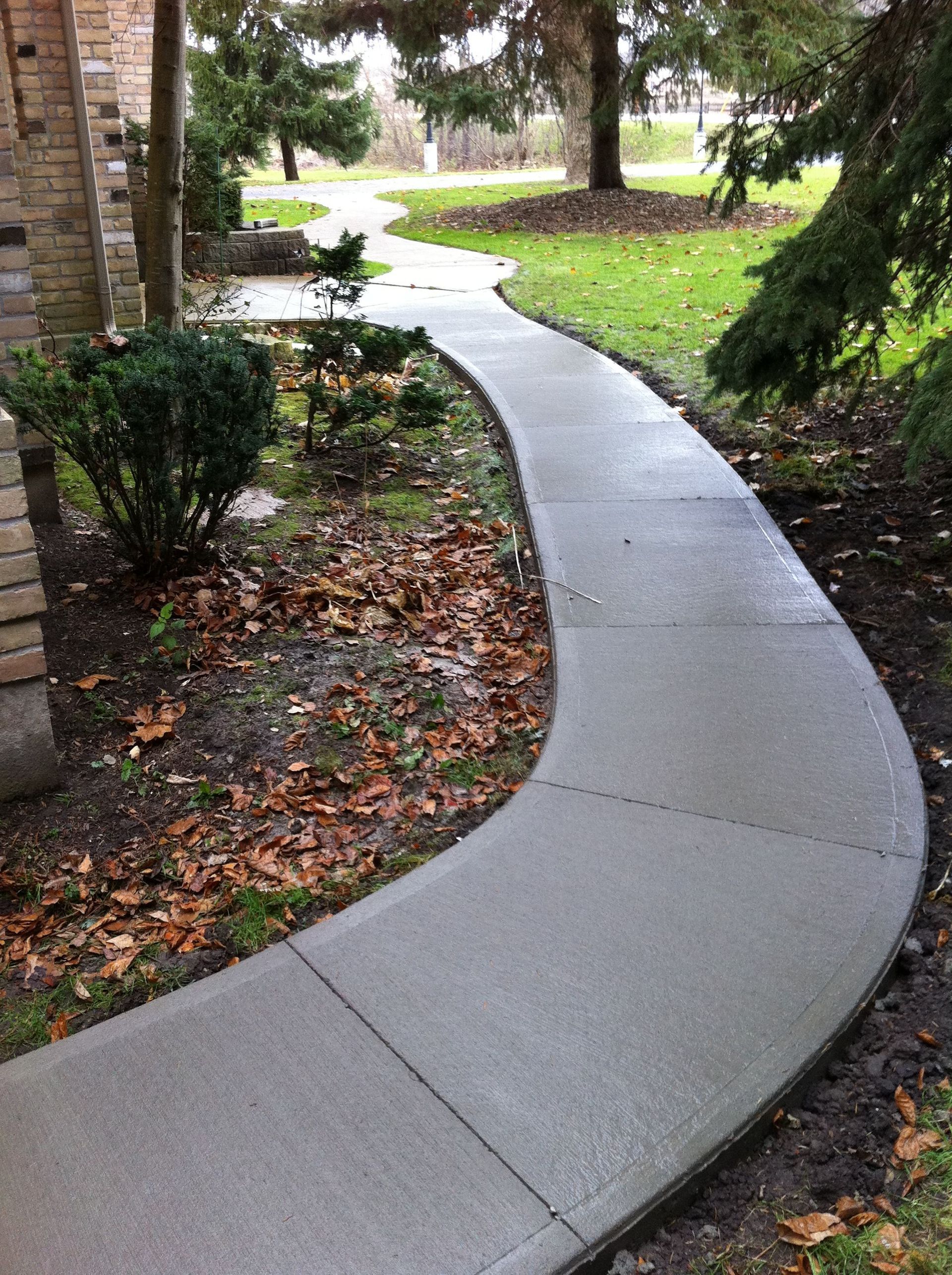 Curving concrete walkway through a yard, bordered by greenery and a brick building.