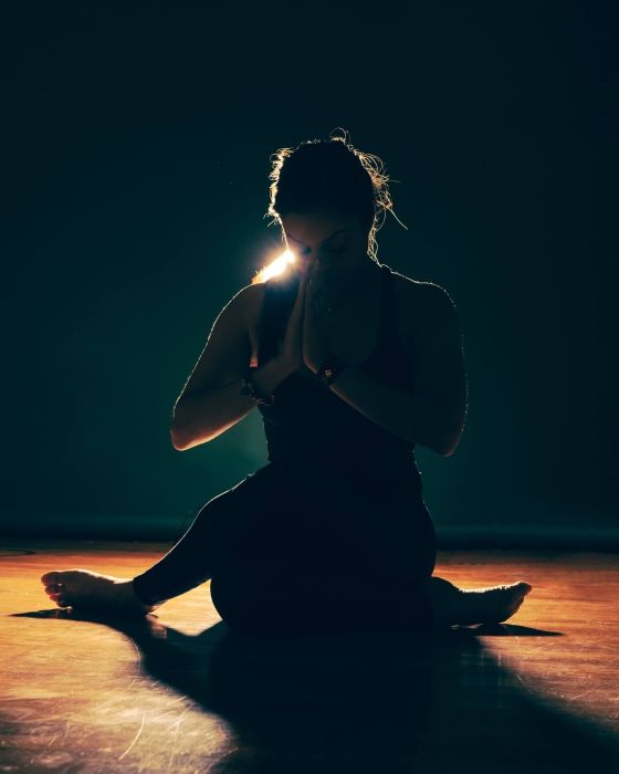A woman is sitting on the floor in a dark room with her hands folded in prayer.