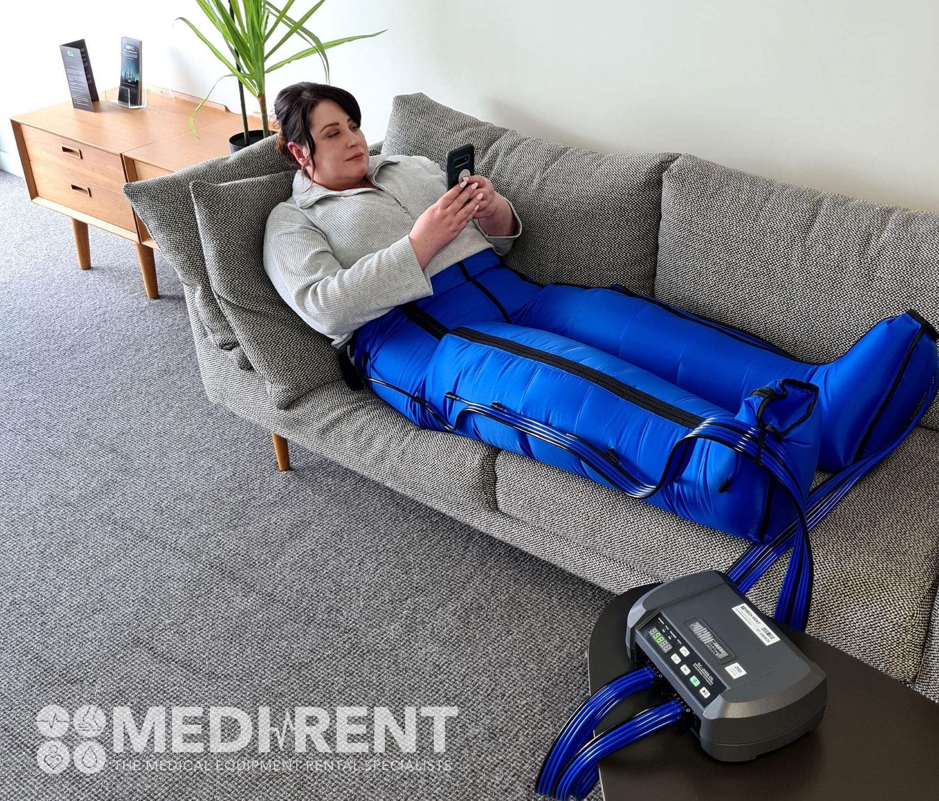 Woman using a leg compression device on a couch, looking at a phone. The device and legs are blue.