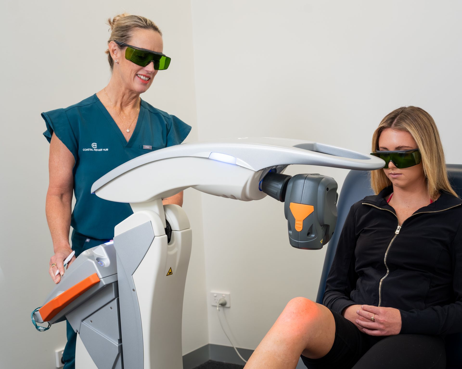 Doctor operating a laser therapy machine on a patient's knee in a clinic. Both wear protective glasses.