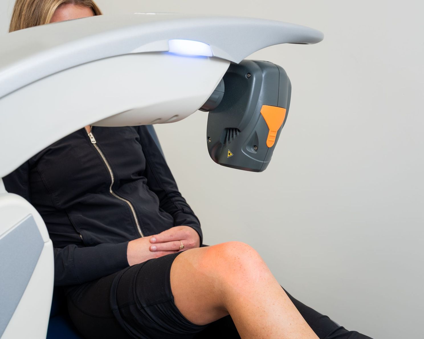 Woman receiving knee treatment from medical device; orange and gray colors.
