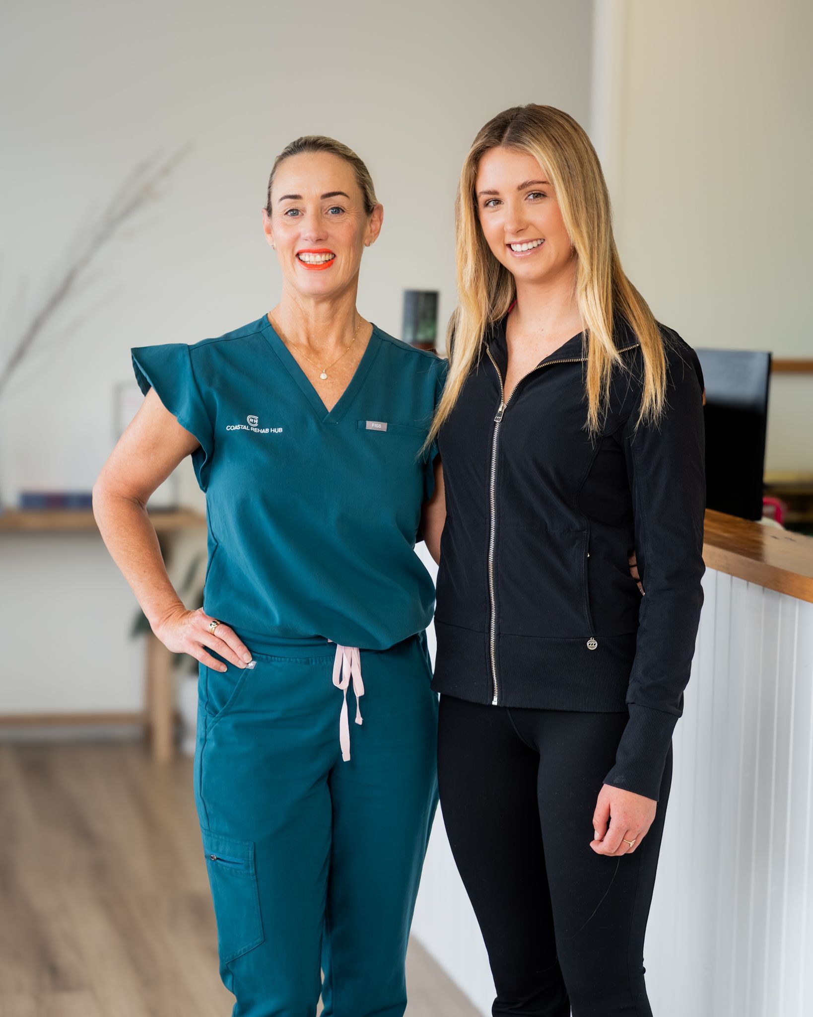 Two women standing indoors, one in teal scrubs and one in black, smiling at the camera.
