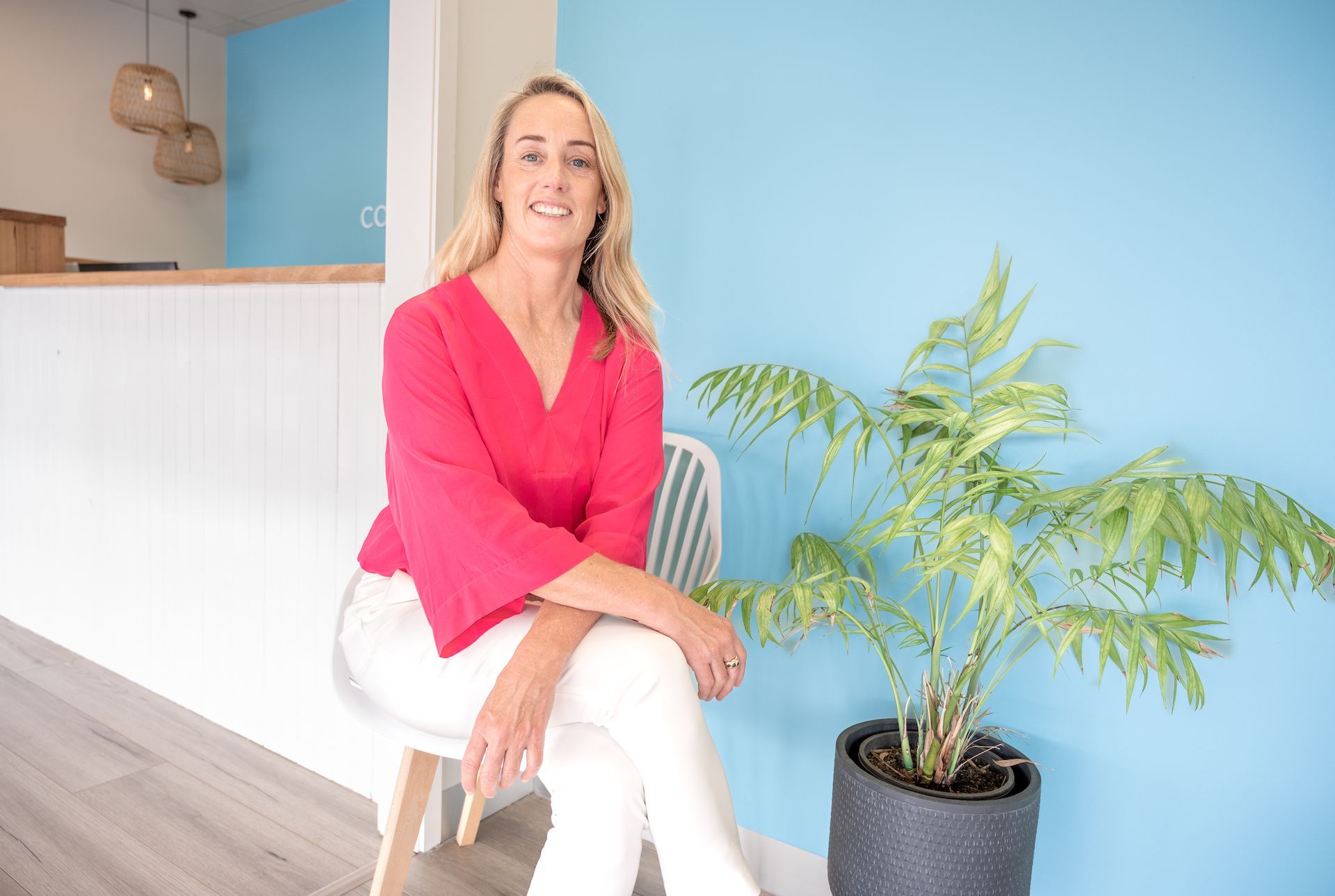 Woman in pink top and white pants sitting on a white chair next to a potted plant, with a blue wall backdrop.
