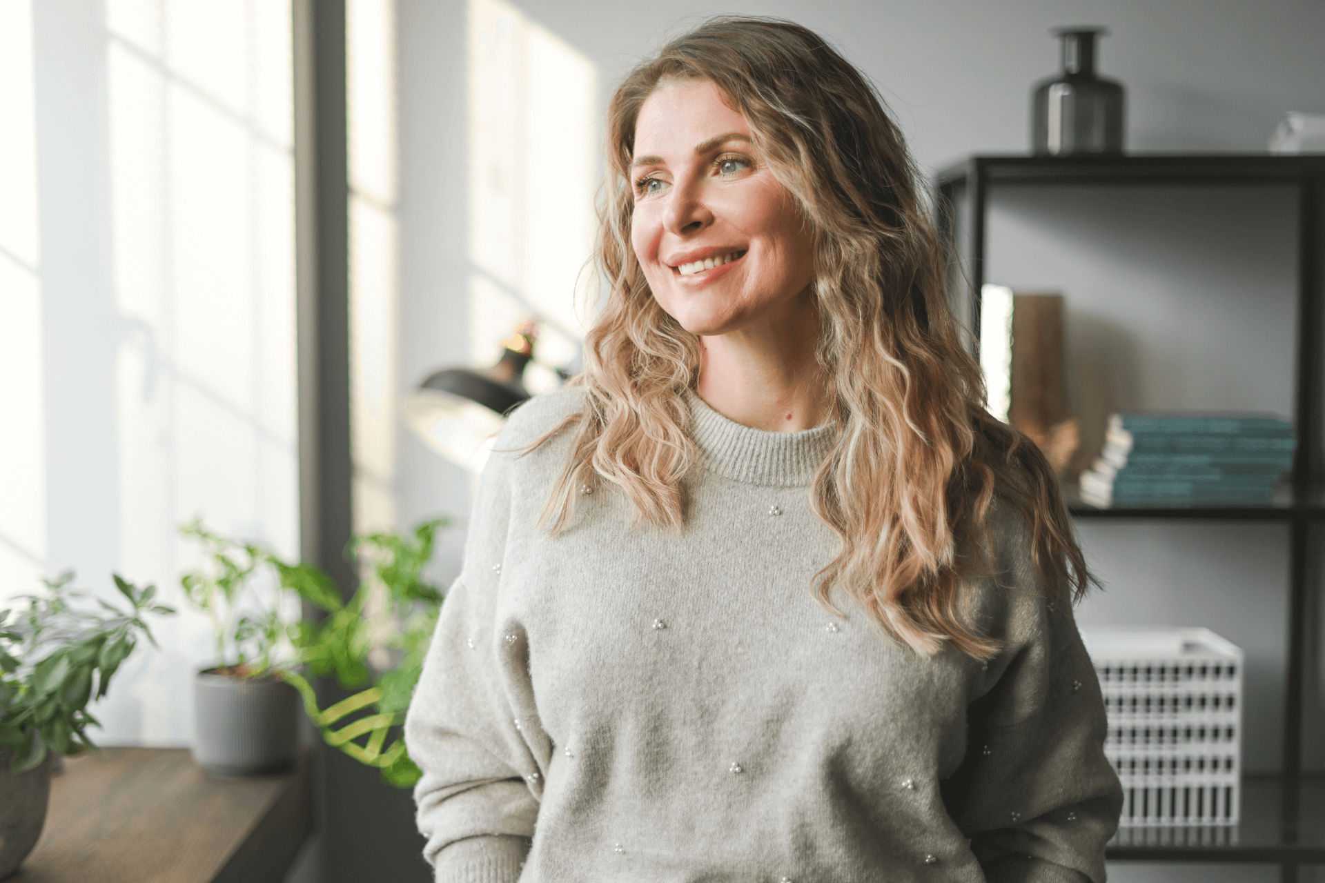 Woman smiling, looking out window.  Blond hair, gray sweater, standing in sunlit room with plants and shelving.