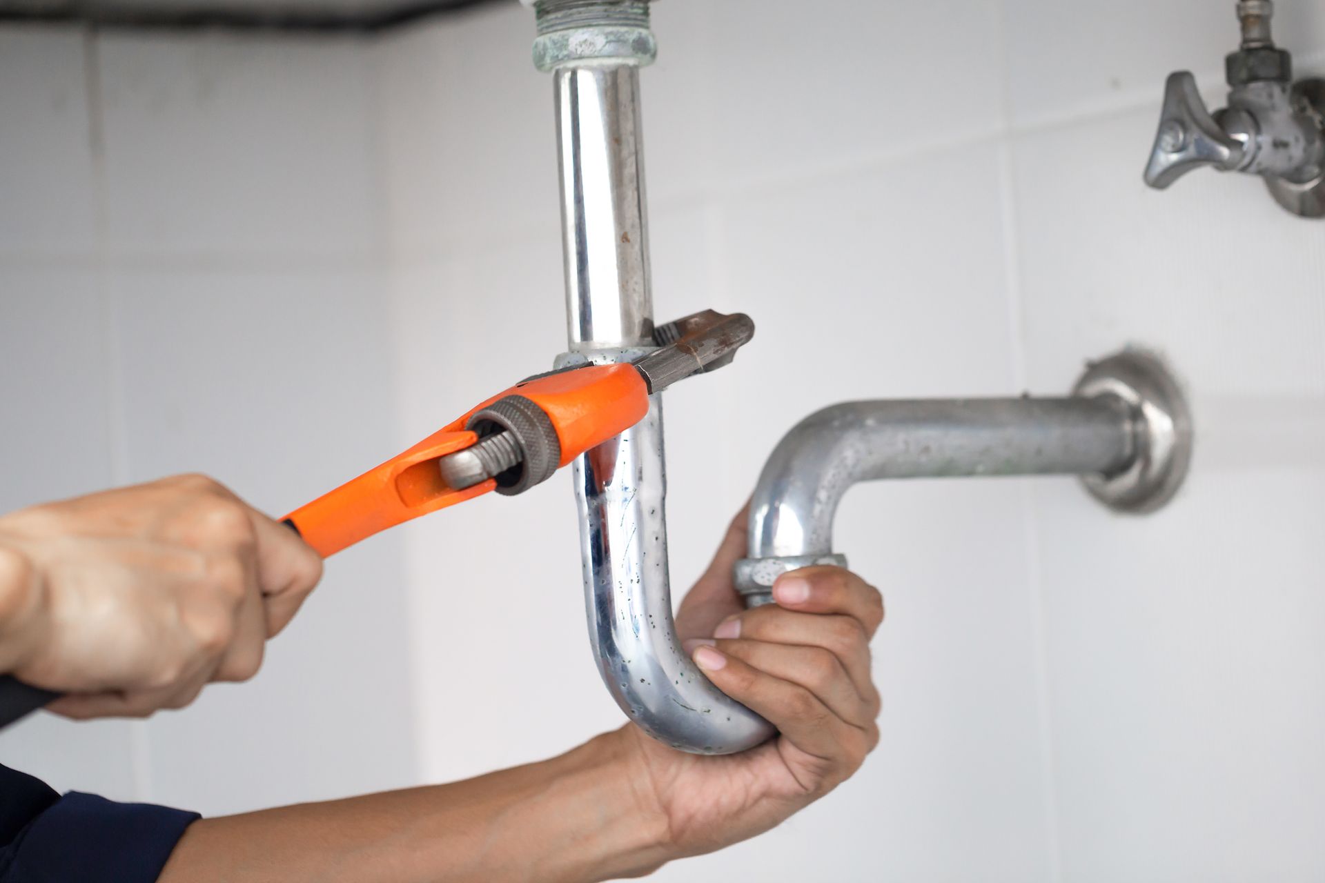 Licensed plumber tightening chrome drain pipes with an orange pipe wrench under a home sink. Licensed plumber tightening chrome drain pipes with an orange pipe wrench under a home sink.