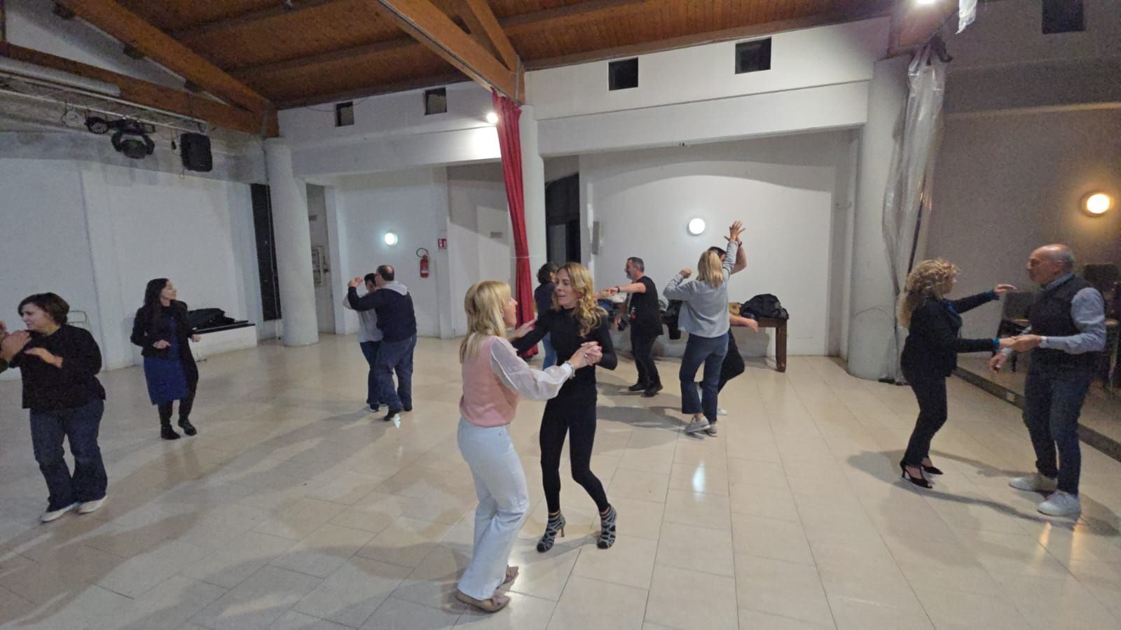 Persone che ballano in una spaziosa sala con travi in legno. Alcuni ballerini sono in coppia, altri da soli.