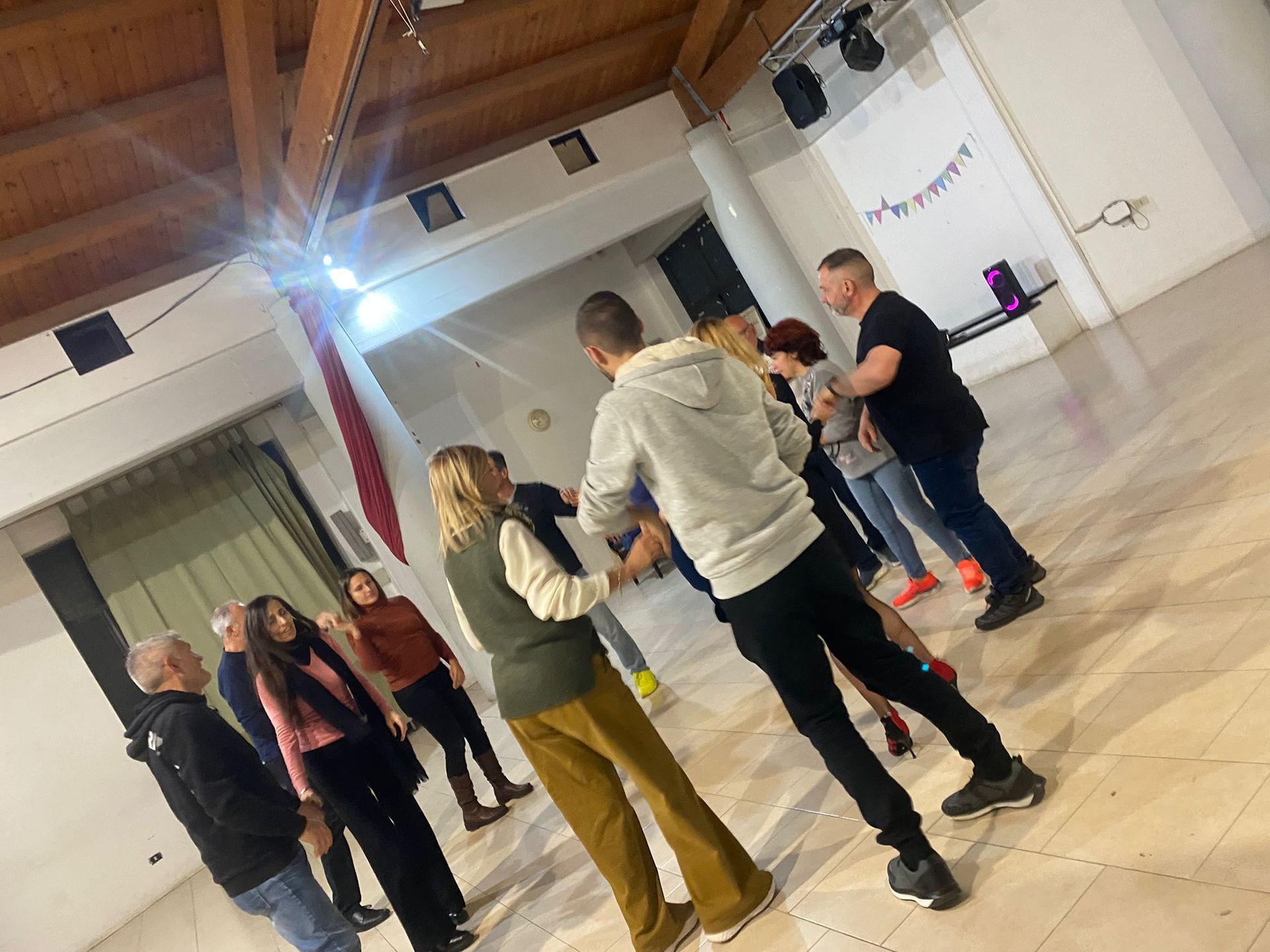 Persone che ballano in una sala da ballo illuminata. Alcuni si tengono per mano e provano i passi. Travi in legno e pareti bianche.